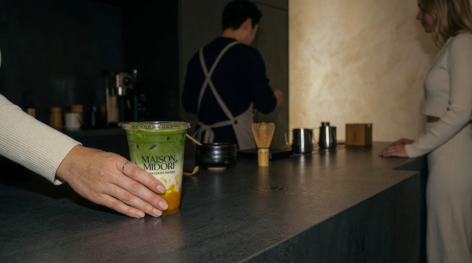 Gast hält einen geschichteten Iced Matcha im Maison-Midori-Becher auf der dunklen Theke, während im Hintergrund ein Barista in Schürze Matcha zubereitet.