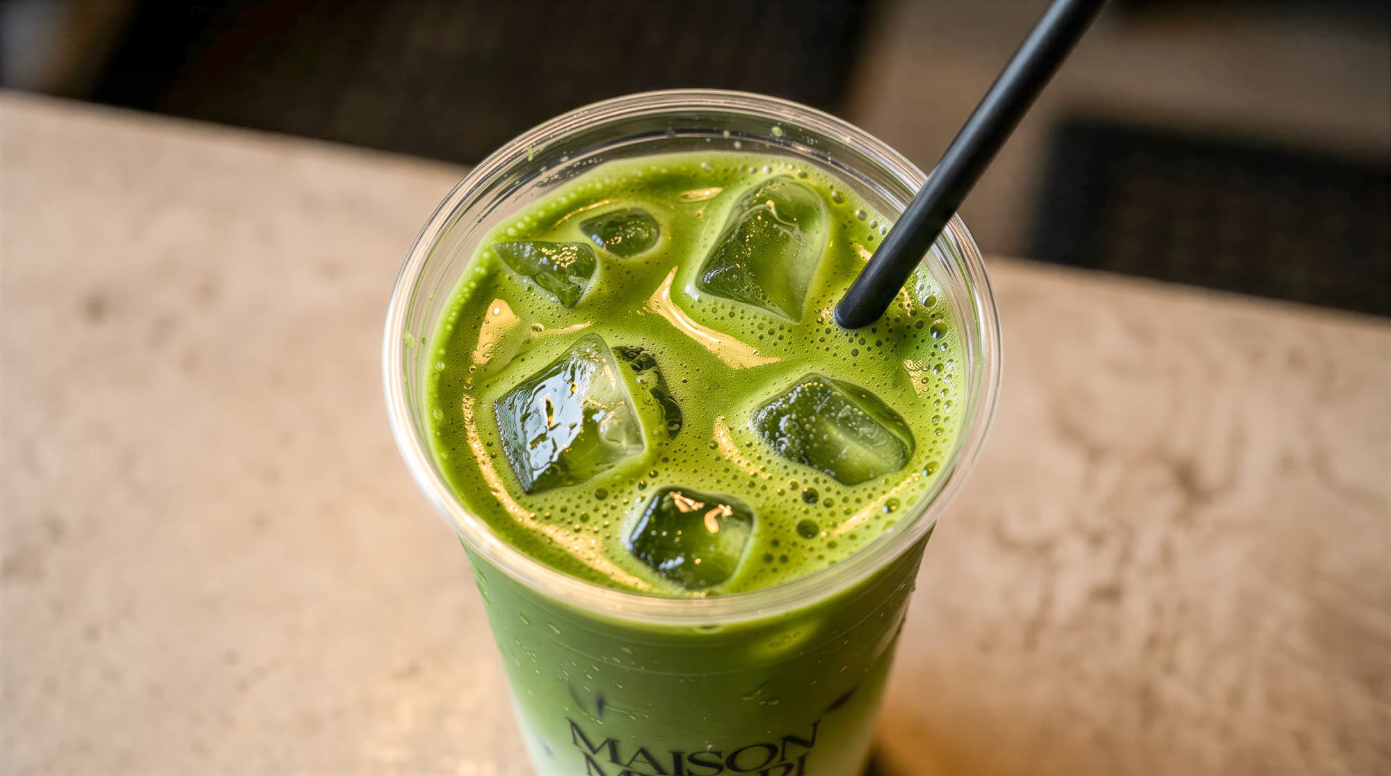 Nahaufnahme eines Iced Matcha im Maison-Midori-Becher mit Eiswürfeln und schwarzem Strohhalm