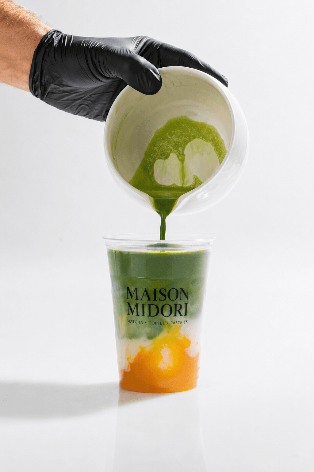 Barista mit schwarzem Handschuh gießt frisch gewhiskten Matcha aus einem Krug in einen Maison-Midori-Becher mit Mango-Schicht