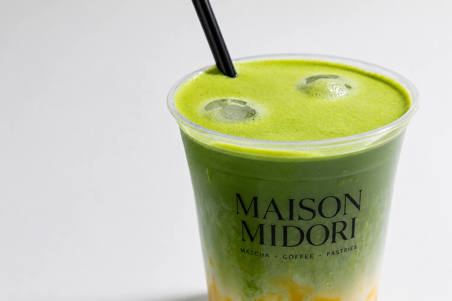 Nahaufnahme von oben auf einen frisch gegossenen Iced Matcha im Maison-Midori-Becher mit schwarzem Strohhalm