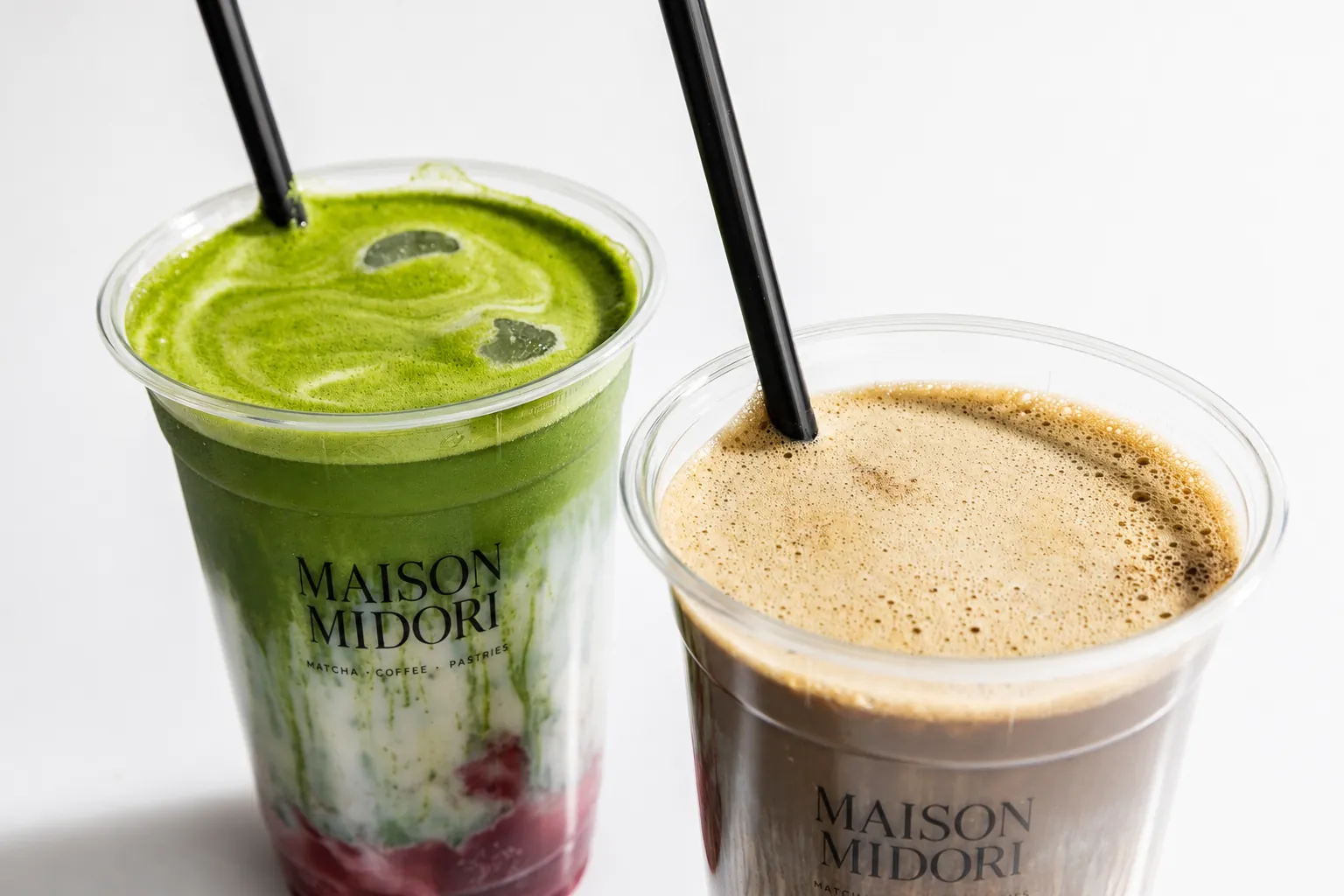 Zwei Maison-Midori-Becher nebeneinander: links ein geschichteter Erdbeer-Matcha, rechts ein cremiger Iced Coffee, beide mit schwarzem Strohhalm