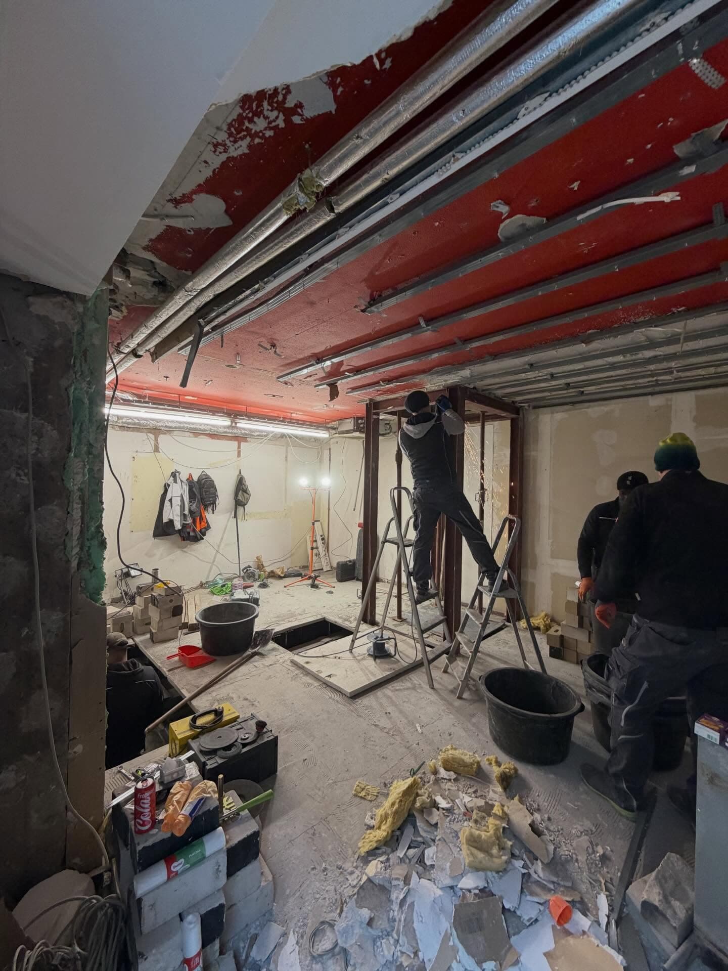 Das Ladenlokal in der Gangolfstraße im Rohzustand, Anfang 2026, drei Bauarbeiter beim Schweißen einer Stahltür-Zarge zwischen freigelegter Decke mit roten Stahlträgern, abgeschlagenem Putz und Bauschutt, kurz bevor Maison Midori entstand.